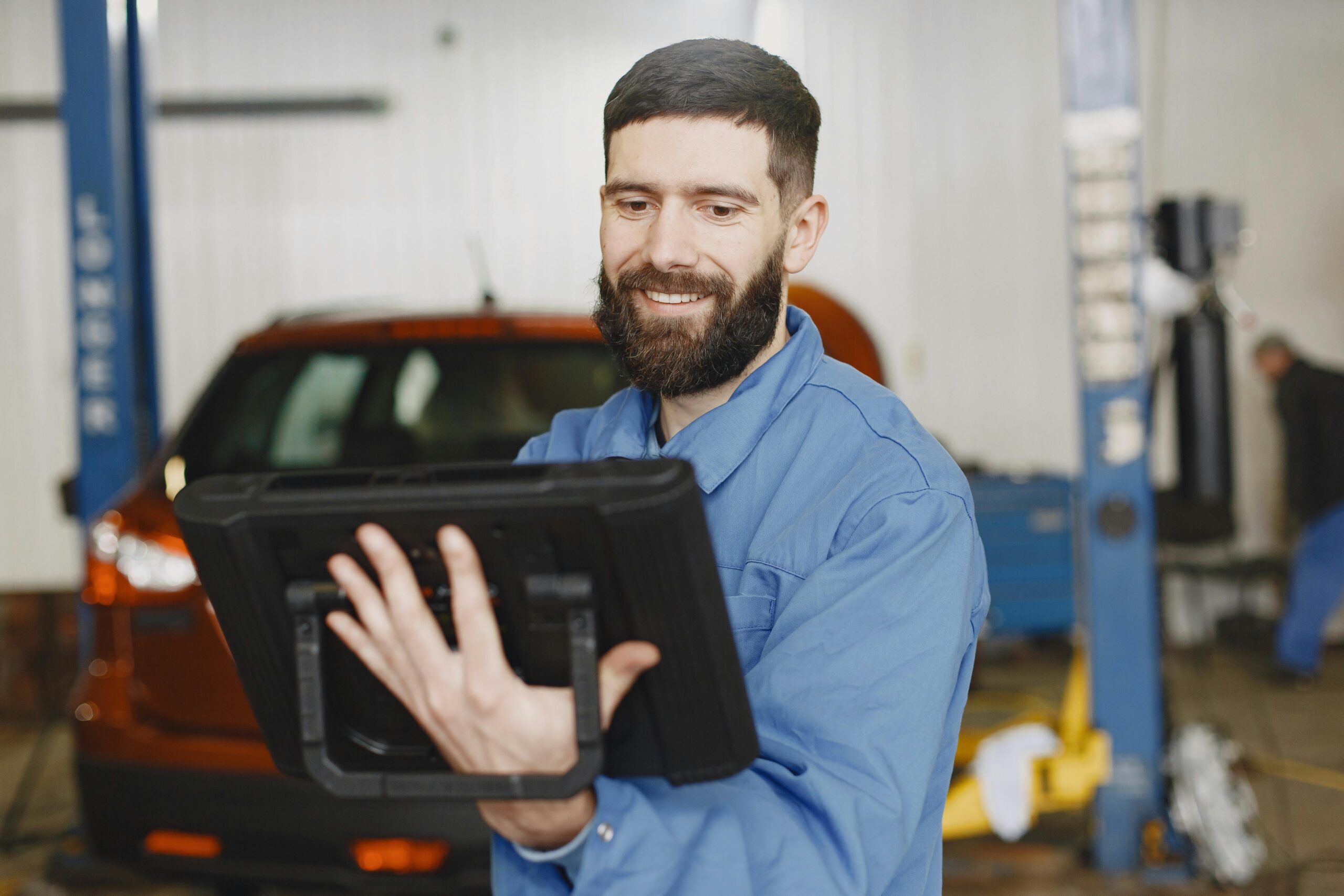 Smiling mechanic in blue uniform uses a digital tablet for vehicle diagnostics in a garage.